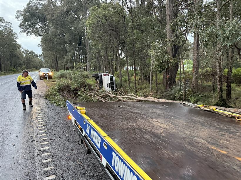 Tilt tray lowered beside a wet highway with a rolled vehicle in the trees